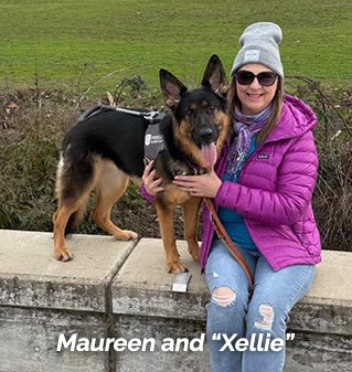 Mareen and her German shepherd guide dog, 'Xellie' sit on a cement wall.  Maureen wears sunglasses, a pink puffy jacket, a gray beanie and a blue sweatshirt under the jacket and a pair of jeans.  Xellie stands next to her on the cement seat garden wall barrier in a harness with head slightly tilted to the side and ears alert.